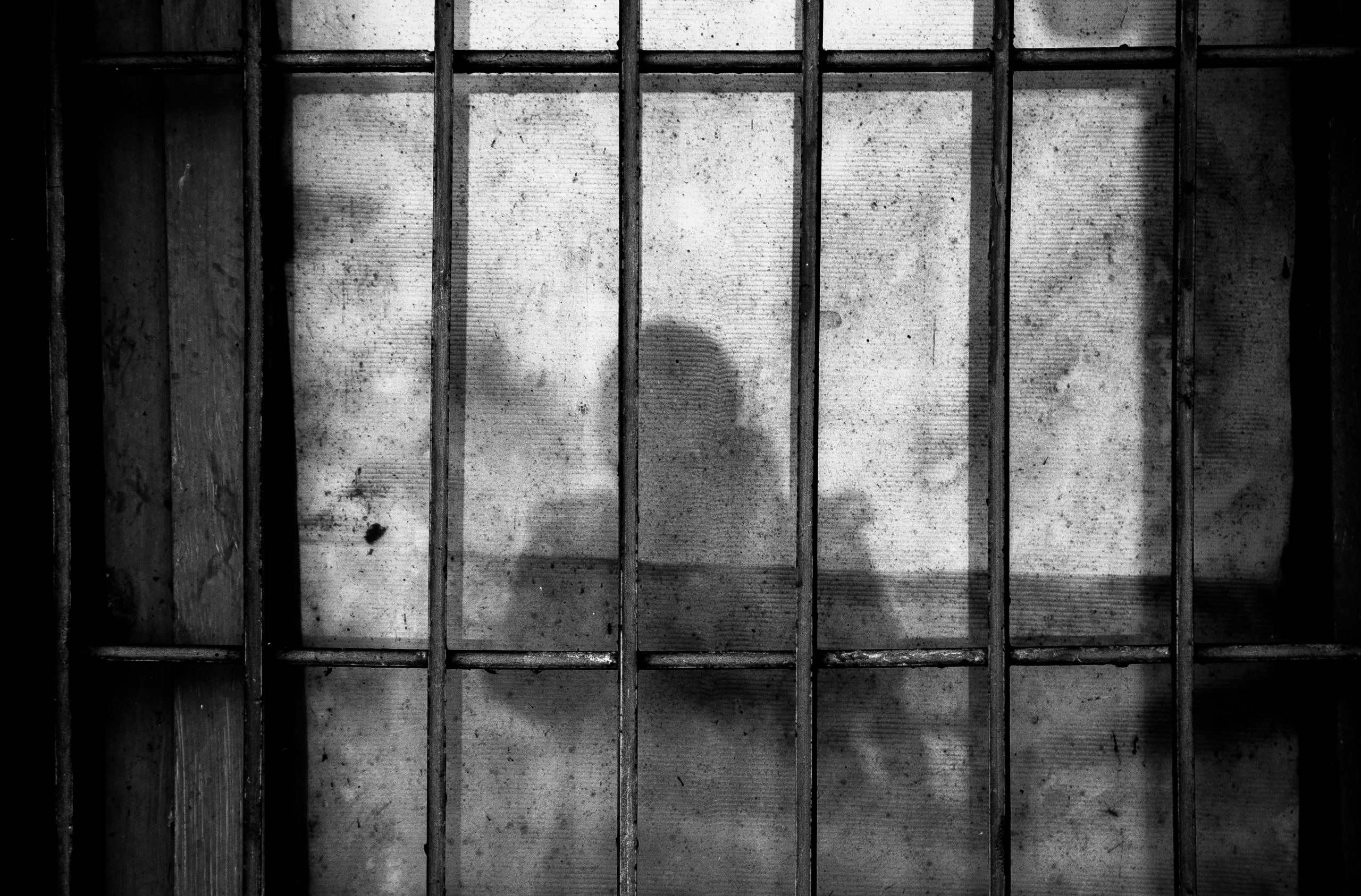 Black and white image depicts the blurred silhouette of a person behind a barred window