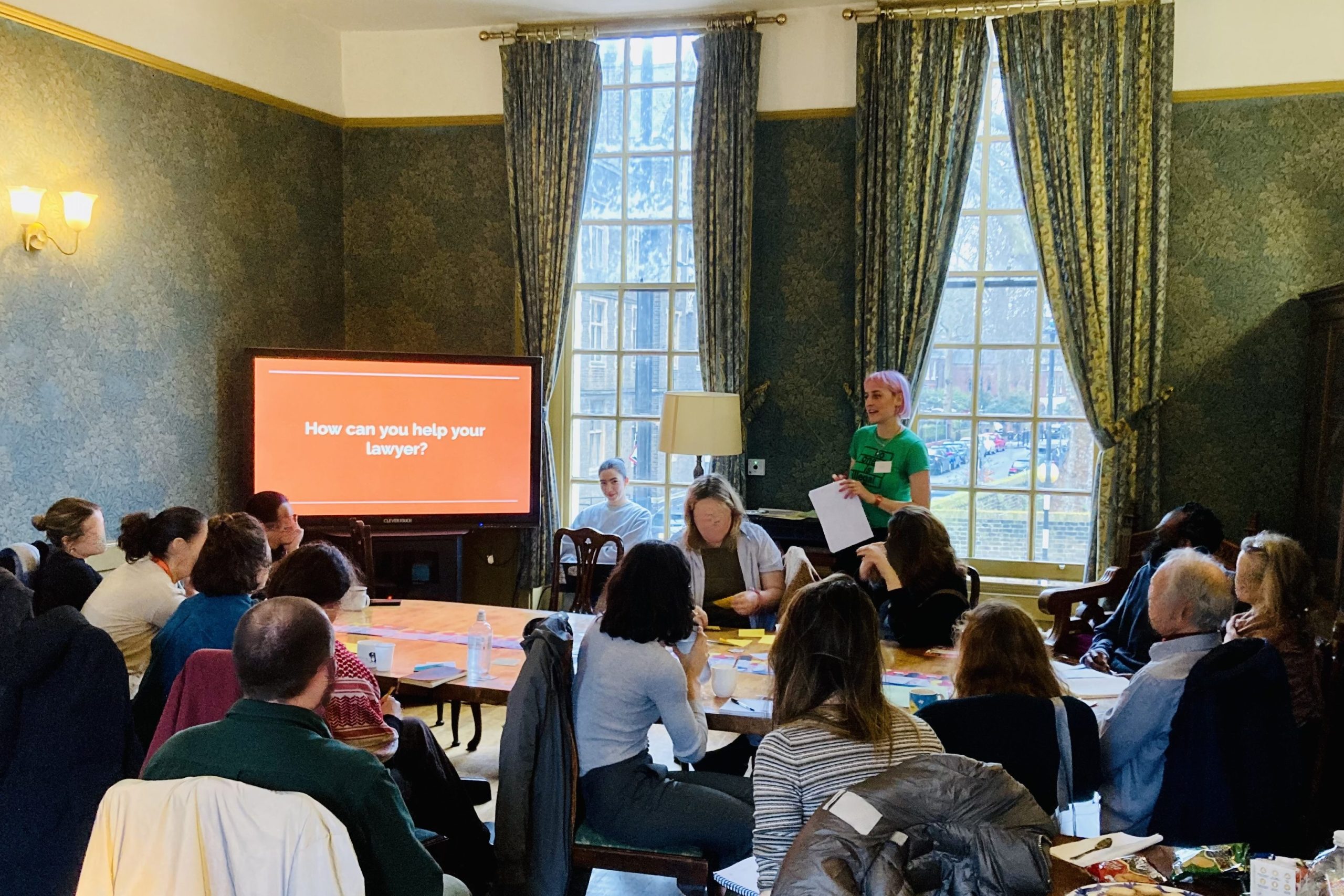 a room full of people sat around a table, watching a presentation on 'how can you help your lawyer?'