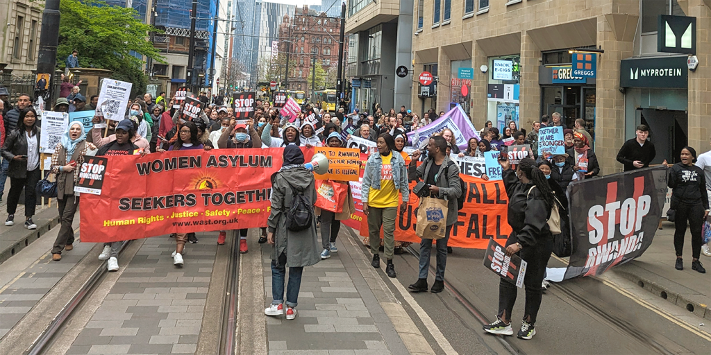 These Walls Must Fall campaigners and allies marching against the Rwanda plan in central Manchester. Some hold banners.