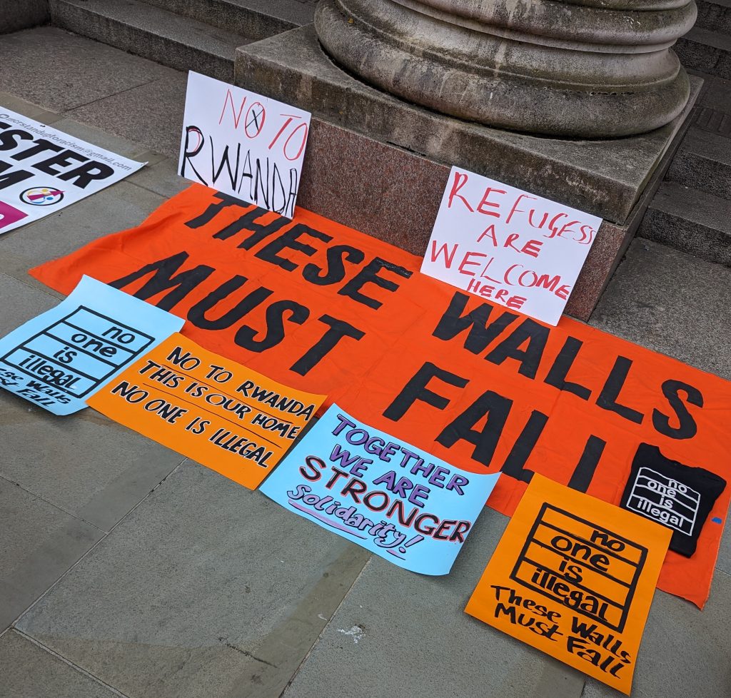 Placards used at a protest, laid on the ground. The placards are different colours (orange, blue, and white) and feature slogans like 'These Walls Must Fall', 'No to Rwanda!' and 'No One Is Illegal'