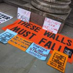 Placards used at a protest, laid on the ground. The placards are different colours (orange, blue, and white) and feature slogans like 'These Walls Must Fall', 'No to Rwanda!' and 'No One Is Illegal'