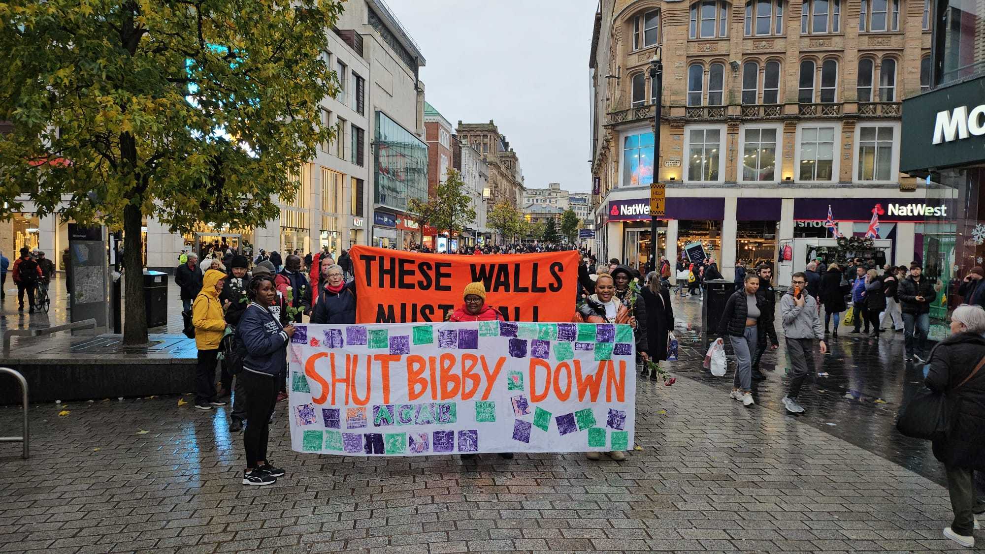 These Walls Must Fall campaigners holding up banners at a protest. One says 'These Walls Must Fall' and the other says 'Shut Bibby Down'