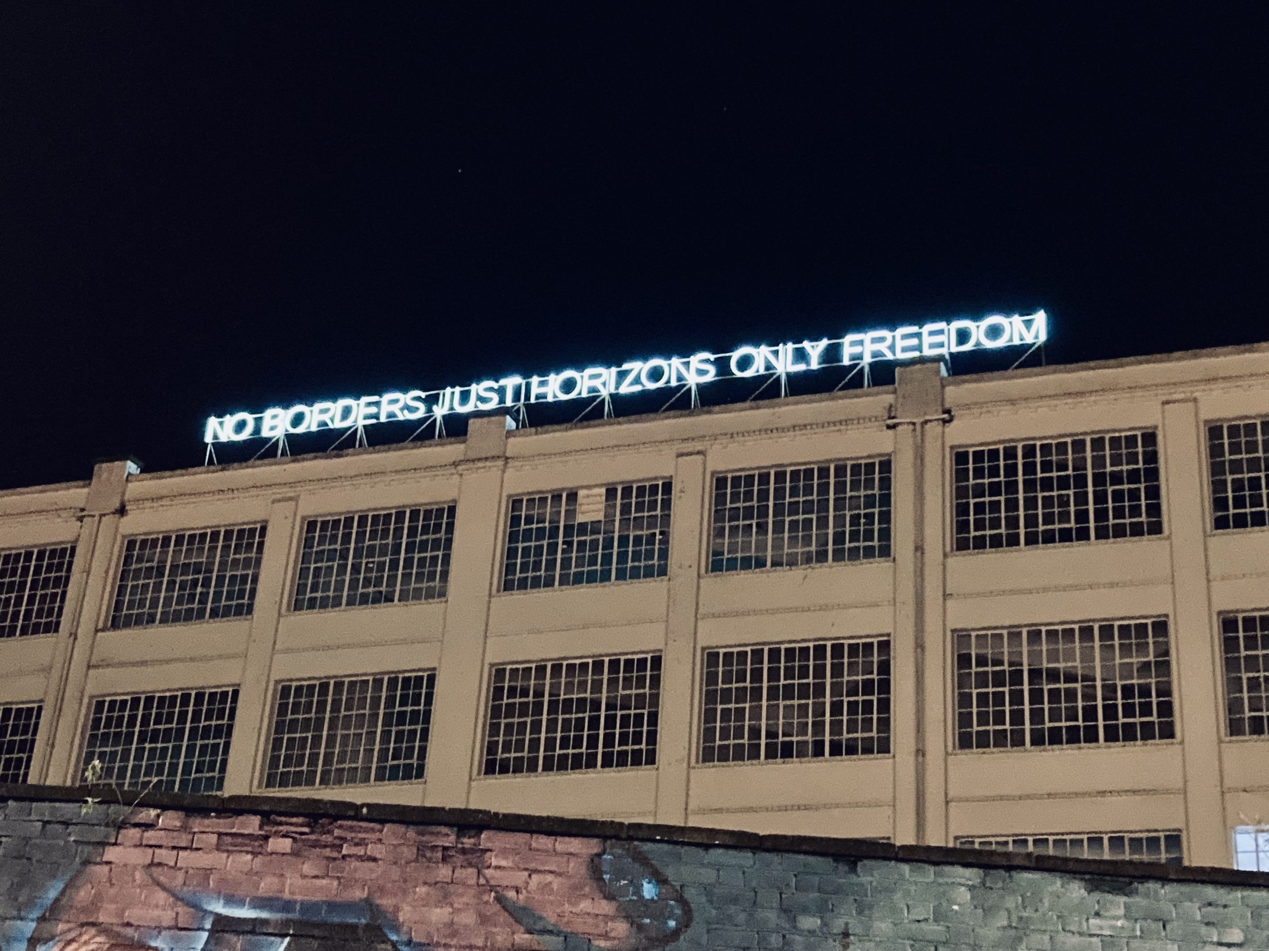 a fluorescent light sign atop a building that reads: NO BORDERS JUST HORIZONS ONLY FREEDOM